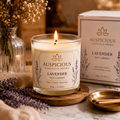 A lit Auspicious Lavender Soy Candle in a clear glass jar sits on a wooden coaster, with the product box behind it and dried lavender sprigs and a glass vase in a warm, styled setting.