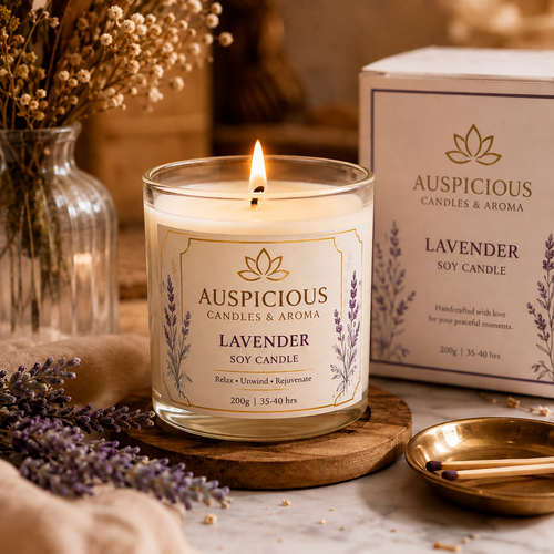 A lit Auspicious Lavender Soy Candle in a clear glass jar sits on a wooden coaster, with the product box behind it and dried lavender sprigs and a glass vase in a warm, styled setting.
